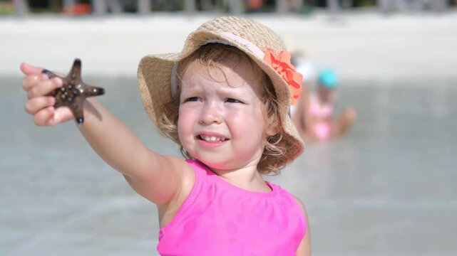 A cheerful baby girl joyfully holds a starfish in her hand while relaxing on a warm tropical beach with her family, slow motion. Vaction and travel concept