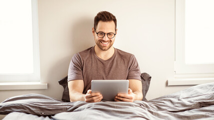 Man Using Digital Tablet Watching Movie Online Sitting In Bed In Bedroom At Home On Weekend. Guy Relaxing Browsing Internet On Computer. Gadgets Lifestyle Concept. Panorama
