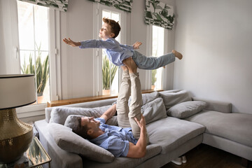 Strong young father lying on sofa in living room, lifting his little 8s son into air as he flying,...