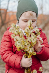 Young child holding a bouquet of fluffy willow branches in early spring, wearing a red jacket and green hat, standing outdoors in a natural rural setting with a blurred background.