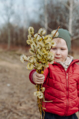 Young child holding a bouquet of fluffy willow branches in early spring, wearing a red jacket and green hat, standing outdoors in a natural rural setting with a blurred background.