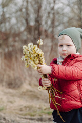 Young child holding a bouquet of fluffy willow branches in early spring, wearing a red jacket and green hat, standing outdoors in a natural rural setting with a blurred background.