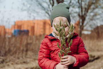 Young child holding a bouquet of fluffy willow branches in early spring, wearing a red jacket and green hat, standing outdoors in a natural rural setting with a blurred background.