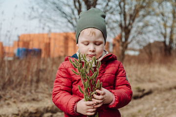 Young child holding a bouquet of fluffy willow branches in early spring, wearing a red jacket and green hat, standing outdoors in a natural rural setting with a blurred background.