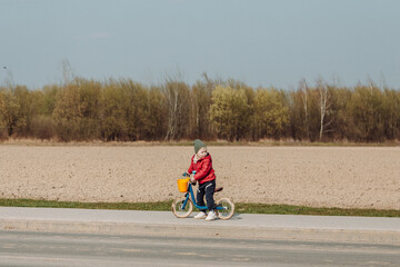 A child in a red jacket and green hat rides a blue balance bike on a paved path next to a field. The child is facing away from the camera, and the field in the background is brown and barren.