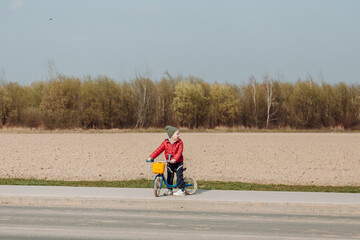 A child in a red jacket and green hat rides a blue balance bike on a paved path next to a field. The child is facing away from the camera, and the field in the background is brown and barren.