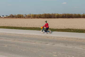A child in a red jacket and green hat rides a blue balance bike on a paved path next to a field. The child is facing away from the camera, and the field in the background is brown and barren.