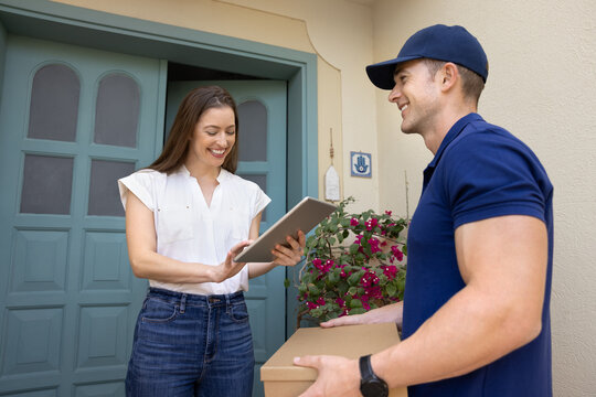 Happy young woman holds tablet, put signature on digital confirmation form while courier holding cardboard box standing together on porch of female client. Professional and reliable doorstep delivery