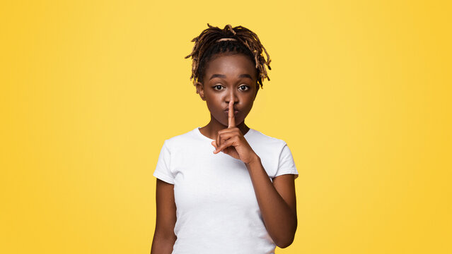 Pretty silent black woman making hush gesture, yellow studio background with copy space. Attractive african american young lady showing silence gesture, touching her lips with one finger
