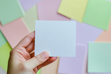 Obraz premium Hand holding blank white square card against pastel background, showcasing a simple yet versatile mockup for text or advertisement
