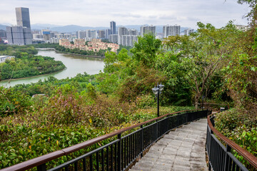 Fototapeta premium A view from a hilltop overlooking a cityscape with a river winding through it. Stone stairs with a metal railing lead down the hill towards a lush green landscape of Sanya, Hainan, China.