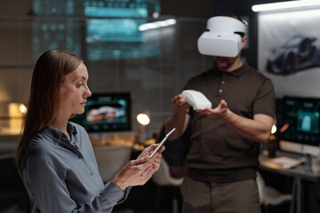 Side view of young serious businesswoman looking at tablet screen while networking against male colleague making virtual presentation