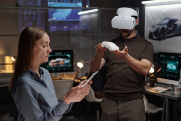 Young male engineer in VR headset holding car model on hands while standing in front of female colleague using tablet