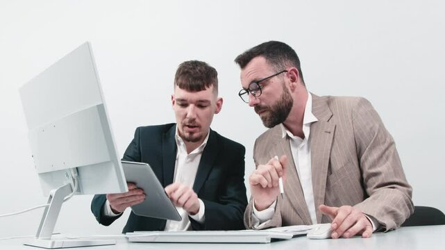 Two business men discuss an online project or presentation at a computer with a tablet. Focused coworkers work in pairs at a computer using apps. The concept of mentoring, teamwork and brainstorming. 