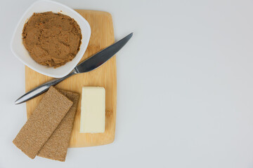 On the left, on a wooden board, there is a white plate with liver pate, slices of bread, butter and a knife on a white background with space for text. The concept of healthy food, a healthy snack