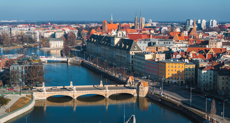 Aerial view of Wroclaw, Poland, showing the Oder River, a stone bridge, Wroclaw University, Wroclaw Cathedral, red rooftops, and a clear blue sky.