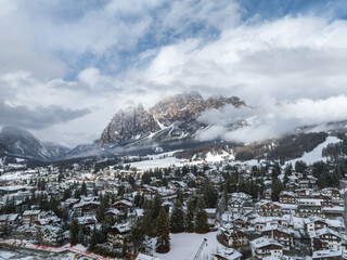 Snow covered rooftops, evergreen trees, and Dolomite peaks surround Cortina d'Ampezzo. Ski slopes and construction for Winter Olympics 2026 are visible.