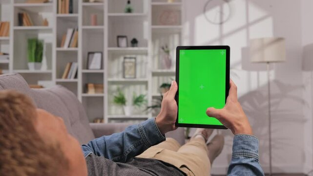 Closeup shot of man lying on sofa using tablet in vertical position with green screen mockup in bright room using different gestures while watching video.