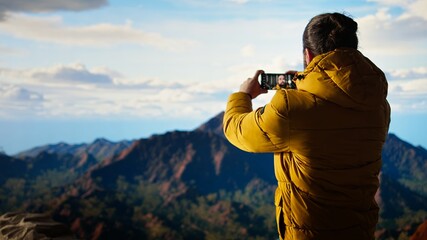 Fototapeta premium Young adventure seeker taking photos on the mountain top using his phone, capturing the moment of his achievement. Nature lover hiker climbing the mountain range and sitting on the peak. Camera A.