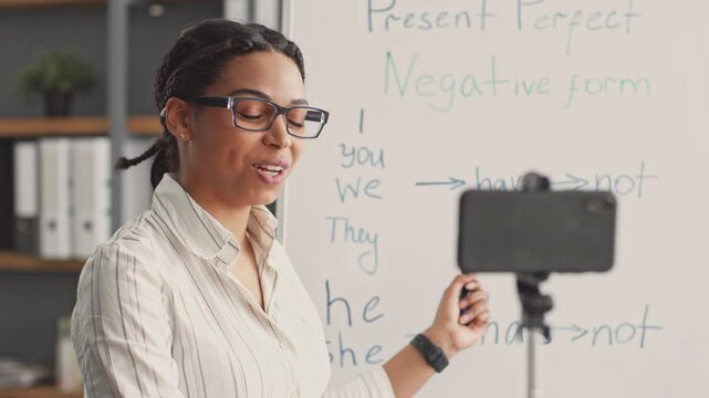 A cheerful African American woman is actively teaching English grammar in an online tutoring session, using a whiteboard to explain the present perfect tense and engaging with her students.