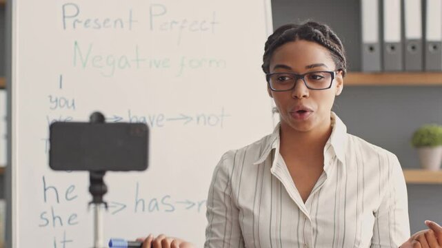 An African American woman conducts an online tutoring session, teaching English grammar. She uses a smartphone to connect with her students while standing by a whiteboard.