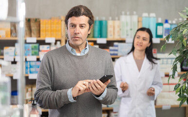 Mature man with a phone in hands flips through an online catalog and looks at the shelves with pills and medicines in a pharmacy. Pharmacist helps the client choose the right medicines