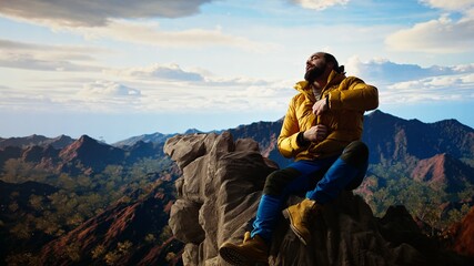 Naklejka premium Young traveler in warm clothing reaches the peak with deep sense of satisfaction, feeling very tired but proud of his achievement. Adventure seeker accomplishing a goal. Camera A.
