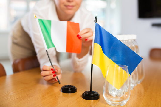 Closeup of national flags of Ukraine and Italy in hands of female office coordinator preparing table in meeting room for international negotiations, selective focus. Concept of diplomatic partnerships