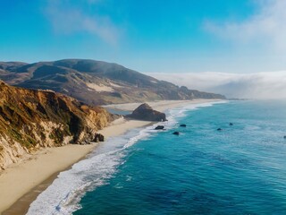 Scenic aerial view of the California coastline in the United States. The Pacific Ocean meets sandy beaches and rocky cliffs under a clear sky. Fog rolls over the hills.