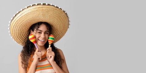 Beautiful young African-American woman in sombrero hat playing maracas on grey background with...