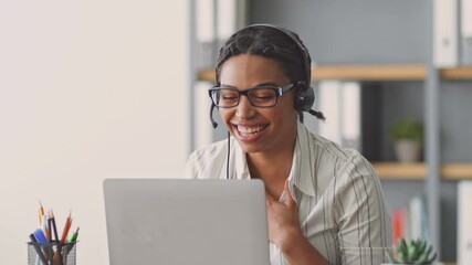 A young African American woman engages with her students during an online class. She wears a headset and smiles while teaching from her bright, organized workspace.