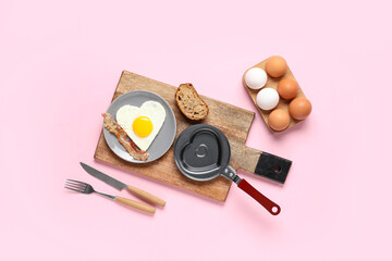 Composition with frying pan, plate with bacon and heart-shaped fried egg on pink background