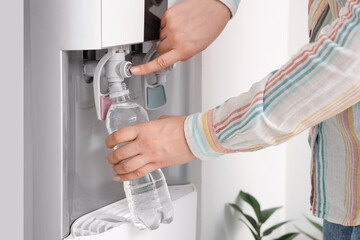 Woman filling plastic bottle with filtered water from cooler near white wall