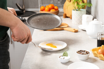 Man with frying pan putting tasty fried eggs onto plate in kitchen, closeup