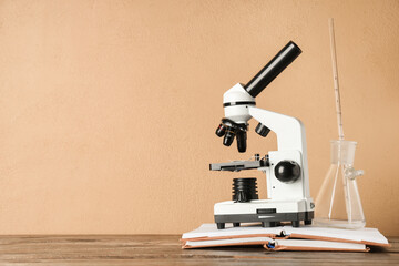 Modern microscope with chemical flask and book on wooden table against beige background