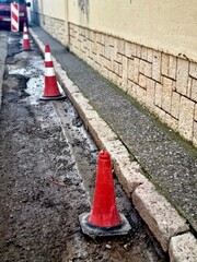 Red and white traffic cones placed along a muddy, damaged roadside near a sidewalk