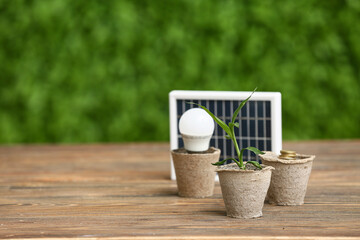 Cups with light bulb, plant and portable solar panel on table outdoors. Green Energy concept