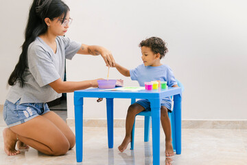 Casually dressed Latino mother and son paint at home with brushes and tempera. The mother supports...