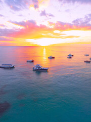 Aerial view of colorful sunrise over ocean in Punta Cana. Boats anchored in calm waters, surrounded by breathtaking scenery. Sky is painted with stunning hues, and gentle waves reflect morning light