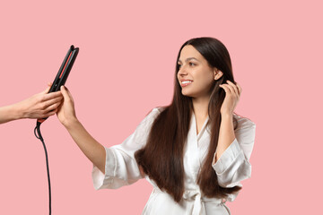 Pretty young woman and female hand with hair straightener on pink background