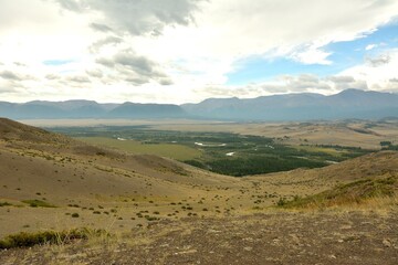 Fototapeta premium View of the endless steppe at the foot of snow-capped mountains through the junction of the foot of two hills on a cloudy summer day.