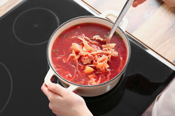 Young woman with cooking pot full of delicious borscht on stove in kitchen, closeup