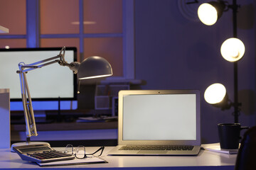Blank laptop with glowing lamp on desk in dark office at night