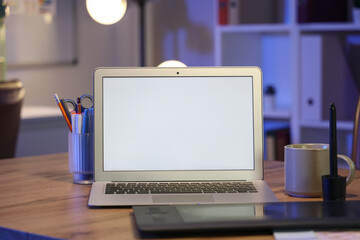Blank laptop with stationery and cup on desk in office at night