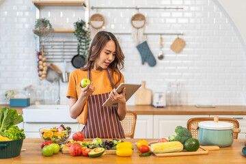 Asian young woman holds a tablet with online audience or simply enjoying the cooking process, health, fruit, freshness, and organic living, digital recipe platform, eating, healthy at kitchen