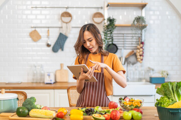Asian young woman holds a tablet with online audience or simply enjoying the cooking process, health, fruit, freshness, and organic living, digital recipe platform, eating, healthy at kitchen