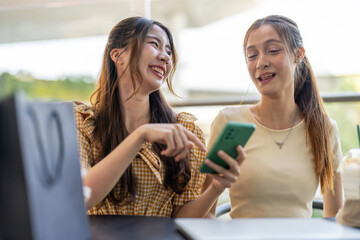 Two diverse women smiling and talking while use phone for online shopping at cafe, searching for deals and discounts with shopping bags on table, concept of e-commerce, retail, digital lifestyle