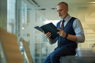 Mature Caucasian businessman in suit sitting on table in board room, looking through papers, copy...