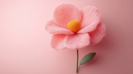 A delicate pink flower made of soft material, featuring a yellow center and green leaf, set against a smooth pink background.