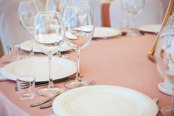 Table setting with light pink tablecloth close up. Wine and beverage glasses, white plates, forks and knives, preparation of the restaurant for the reception of guests